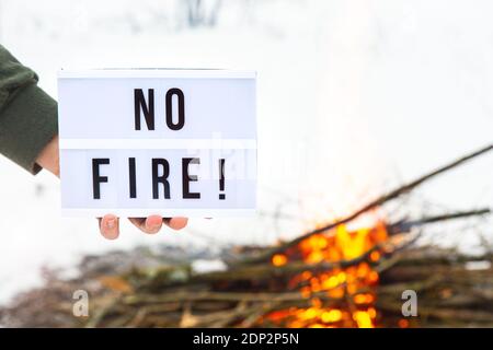 Il concetto di protezione della natura, protezione dagli incendi boschivi, ecologia. Un segno con il testo nessun fuoco nella mano di un uomo su un falò fiammeggiante. Foto Stock