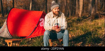 Uomo bearded in camicia a scacchi e cappello seduto su sgabello e bere bevande calde vicino tenda in autunno nella foresta Foto Stock