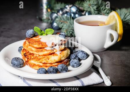 Frittelle fritte fatte in casa con latte condensato e mirtilli sul piatto bianco e una tazza di tè sul tavolo, decorato per Natale. Foto Stock