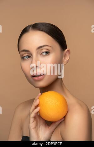 Donna con perfetta pelle del viso, capelli pettinati, che tiene arancione. Ritratto di donna con trucco naturale e agrumi in posa su sfondo beige. Vitam Foto Stock