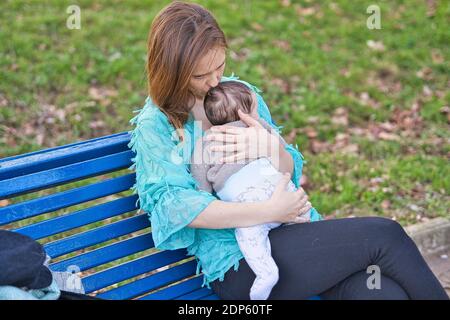 Bella donna che baciava il suo bambino seduto su una panchina in un parco al tramonto. Foto Stock