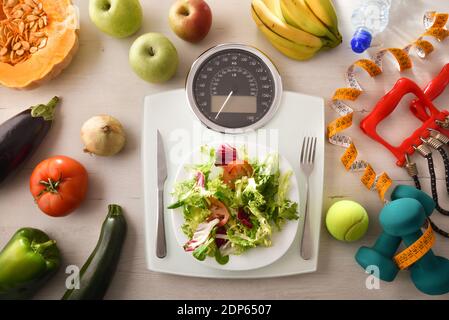 Concetto di controllo del peso con bilancia con piatto per insalate e posate sulla parte superiore e frutta, verdura e accessori sportivi sul tavolo. Vista dall'alto. Orizzontale Foto Stock