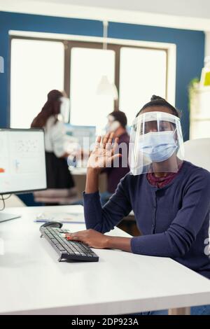 POV di lavoratore africano in chiamata remota sul posto di lavoro seduto alla scrivania indossando maschera contro il covid19. Donna che parla con il team durante la conferenza online mentre i colleghi lavorano in background. Foto Stock
