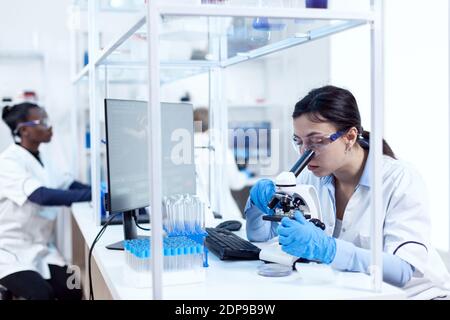 Esperto in genetica facendo ricerca utilizzando il microscopio e assistente africano in background. Camice da laboratorio che indossa una tecnologia moderna durante gli esperimenti scientifici in ambiente sterile. Foto Stock