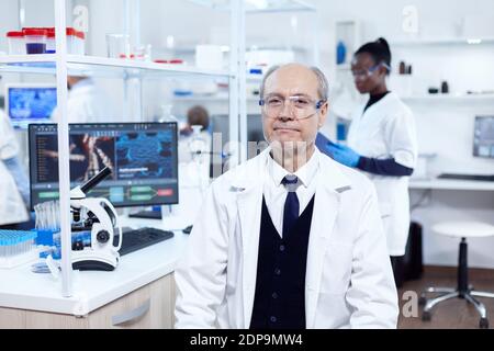 Chimico maschile senior che guarda la fotocamera indossare occhiali protettivi. Anziano scienziato indossando camice da laboratorio lavorando per sviluppare una nuova vacanza medica con assistente africano in background. Foto Stock