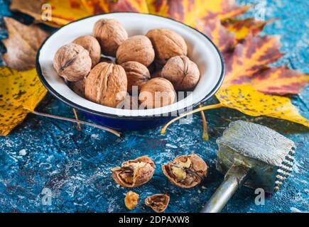 Noci in una ciotola di smalto, noci rotte, martello da cucina, foglie gialle cadute su fondo di cemento blu. Vendemmia autunnale. Foto Stock