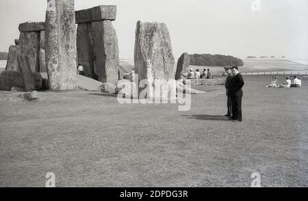 Anni '1950, storico, due uomini in piedi accanto alle antiche pietre di Stonehenge, il famoso momento preistorico sulla piana di Salisbury nel Wiltshire, Inghilterra, Regno Unito. In questo periodo, in un'epoca più rilassata, i visitatori potevano camminare liberamente tra le pietre erette di sarsen senza alcun problema e sedersi su di esse, come stanno facendo le persone, qualcosa che più tardi, quando l'ingresso alle pietre era strettamente controllato, non sarebbe stato permesso. Foto Stock