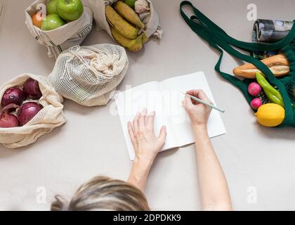 Donna che fa una lista della spesa su un notebook bianco. Molta frutta e verdura sana rimangono all'interno di sacchetti eco-sostenibili. Foto Stock