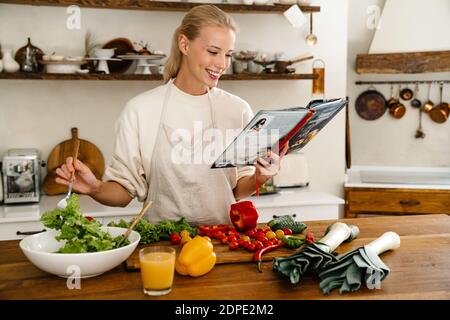 Bella gioiosa donna che legge libro di cucina e sorridendo mentre fa il pranzo in una cucina accogliente Foto Stock