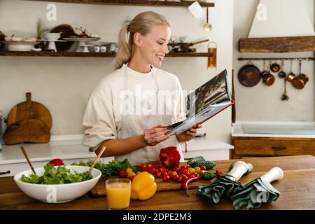 Bella gioiosa donna che legge libro di cucina e sorridendo mentre fa il pranzo in una cucina accogliente Foto Stock