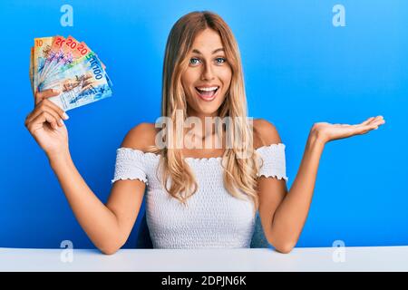 Bella giovane donna bionda che tiene banconote in franchi svizzeri per celebrare il successo con sorriso felice e espressione vincente con mano sollevata Foto Stock