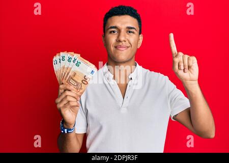 Giovane arabo che detiene 50 banconote in euro sorridendo con un'idea o una domanda che punta il dito con il volto felice, il numero uno Foto Stock