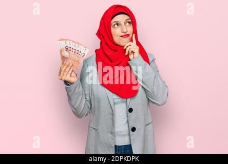 Bella donna ispanica che indossa hijab islamico tenere 10 euro banconote faccia seria pensare alla domanda con la mano sul mento, pensieroso circa con Foto Stock