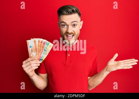 Giovane redhead che tiene banconote di peso filippino che celebra il successo con buon sorriso e espressione vincente con mano sollevata Foto Stock