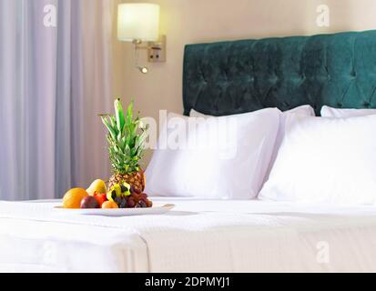 Omaggio di benvenuto fruttato nell'interno dell'hotel sfocato sfondo. Primo piano di sano piatto di frutta prima colazione con ananas su biancheria da letto bianca in camera da letto moderna. Foto Stock