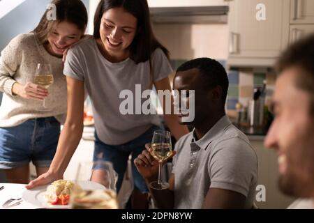 Felici giovani donne che si appoggiano al tavolo mentre si serve la cena fidanzati multietnici durante la festa del vino a casa Foto Stock