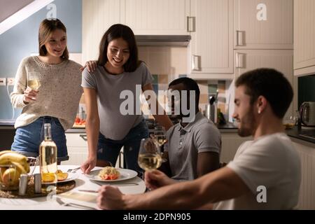 Giovani donne ottimiste sorridendo e servendo piatti gustosi per multirazziale uomini con vino mentre si cena a casa insieme Foto Stock