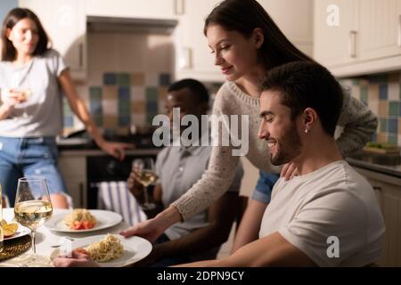 Felice giovane femmina putting piatto con piatto gustoso vicino allegro fidanzato durante la festa di casa con amici diversi
