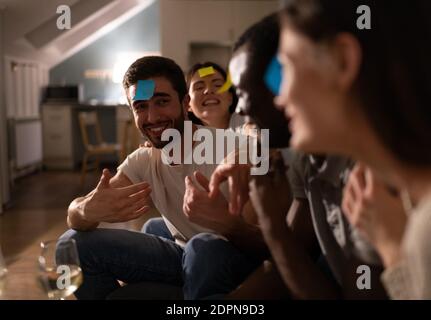 Giovane maschio sconcertato con nota appiccicosa sulla fronte gesturing e. cercando di indovinare chi è lui mentre si gioca divertente gioco con amici diversi durante la festa Foto Stock