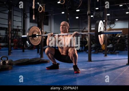 Un potente maschio senior a lunghezza intera che fa squat anteriore come parte di esercizio pulito e a scatti durante un allenamento intenso in palestra Foto Stock