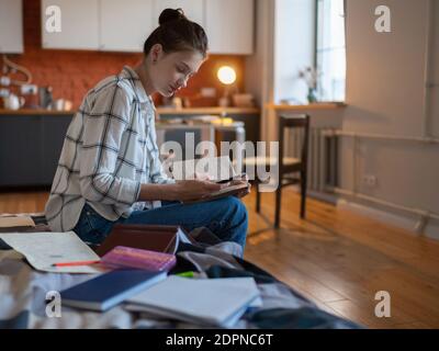 Attraente studentessa adolescente focalizzata utilizzando smartphone e scrivere note per l'esame mentre si è seduti a letto Foto Stock