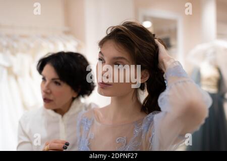 Giovane donna che indossa abito da sposa blu tenendo i capelli e pensando su acconciatura mentre consulente femminile di nozze che dà il consiglio nel matrimonio atelier Foto Stock