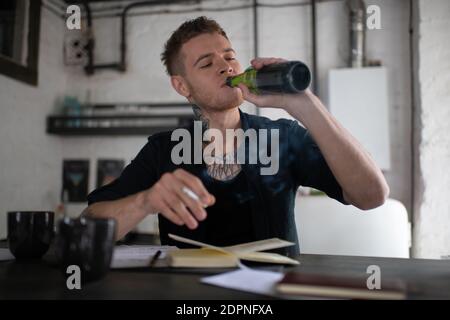 Uomo giovane tatuato che beve birra dalla bottiglia e fuma sigaretta mentre scrivendo al taccuino e sedendosi dal tavolo a. loft urbano Foto Stock