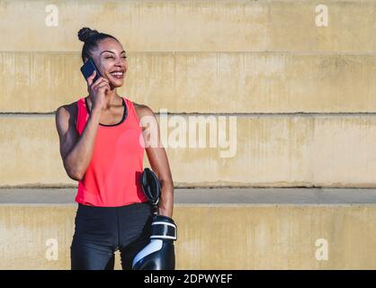 afroamericana attraente giovane donna vestendo abiti sportivi e guanti di boxe in piedi in un parco e parlando al telefono mentre sorride. Spazio di copia sul Foto Stock