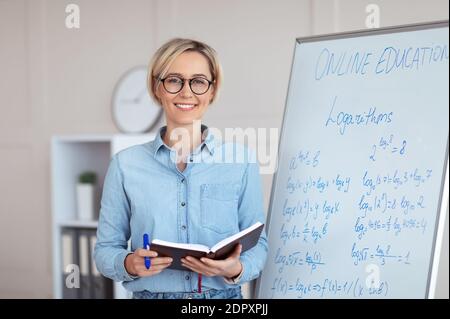 Ritratto di felice giovane insegnante universitario in piedi vicino a lavagna con problemi di matematica, conducendo lezione di internet Foto Stock