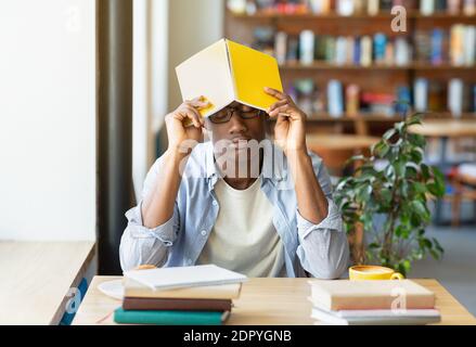 Ragazzo nero overworked che dorme durante i suoi studi al caffè della città, mettendo il libro sulla sua testa, stanco di fare l'assegnazione domestica Foto Stock