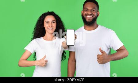 Coppia diversa che mostra lo schermo vuoto dello smartphone gesturing Thumbs-Up, sfondo verde Foto Stock