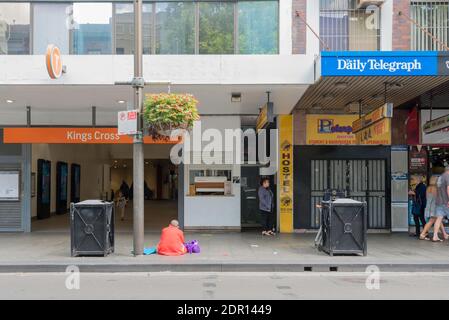 Una persona senza casa si siede sul sentiero tra bidoni spazzatura fuori dalla stazione ferroviaria su Bayswater Road, Kings Cross, Sydney, Australia Foto Stock