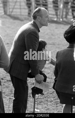 Regno Unito, Inghilterra, Devonshire, Buckfastleigh, 1972. Le gare Point-to-Point si sono svolte a Dean Court sulle paludi Dean, vicino alla A38 tra Plymouth ed Exeter. Uno spettatore con un bambino seduto su un bastone di tiro. Sta fumando una sigaretta e tenendo una scheda o un racebook, un programma di eventi. Foto Stock