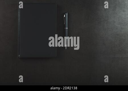 Blocco note nero con una penna nera su sfondo nero. Vista dall'alto, concetto minimalista. Spazio libero. Foto Stock