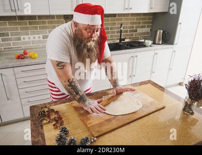 Uomo divertente che indossa la cucina di cappello di Santa in cucina a casa. Buon Natale e Felice Anno Nuovo. Cucina e concetto di vacanza Foto Stock