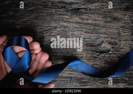 Vista dall'alto della mano maschio che tiene il nastro blu scuro su sfondo di legno con spazio per la copia. Colon, stomaco, cancro colorettale, consapevolezza dell'artrite. Foto Stock