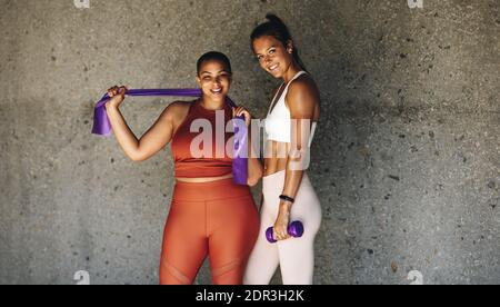 Due donne che si esercitano insieme con la banda di resistenza e dumbbell. Amici femminili in sportswear reggendo la banda di resistenza e dumbbell guardando la macchina fotografica Foto Stock