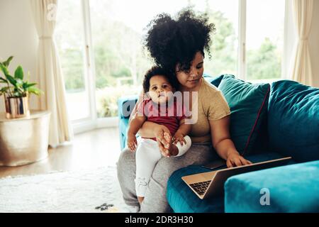 Donna afroamericana bambino seduto su divano utilizzando il computer portatile. Freelance che lavora durante il congedo di maternità. Foto Stock