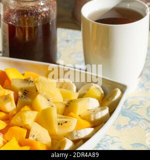 Grande piatto di frutta esotica fresca per colazione, mango, banane, kiwi dorato su un tavolo con tè e marmellata di fragole Foto Stock