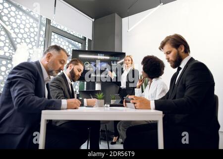 Bella donna d'affari matura bionda motivata intelligente che dimostra infografiche sul digitale di fronte alla sua attività multietnica concentrata Foto Stock