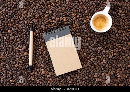 Disposizione piatta con spazio per la copia, (messa a fuoco selettiva) UN taccuino a spirale con una penna e una tazza da caffè bianca con un po' di caffè cremoso vengono posti sui chicchi di caffè. Foto Stock