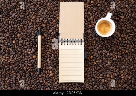 Disposizione piatta con spazio per la copia, (messa a fuoco selettiva) UN taccuino a spirale con una penna e una tazza da caffè bianca con un po' di caffè cremoso vengono posti sui chicchi di caffè. Foto Stock