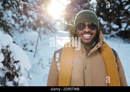 Felice giovane africano in occhiali da sole e caldo abbigliamento invernale in piedi davanti alla macchina fotografica contro i firtrees nella neve durante il freddo nel fine settimana invernale Foto Stock