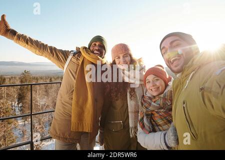 Gruppo di amici interculturali eccitati in winterwear facendo selfie contro sole sul cielo limpido durante la giornata invernale mentre si diverte durante l'escursione Foto Stock
