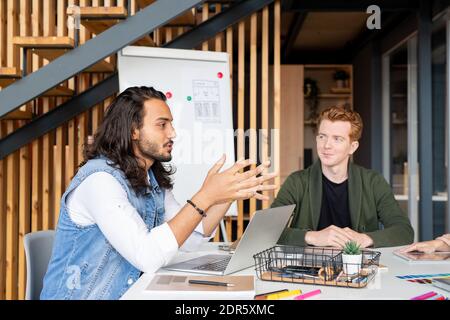 Giovane sviluppatore di app mobile maschile di razza mista che guarda i colleghi durante brainstorming e discussione di idee creative durante la riunione di lavoro Foto Stock