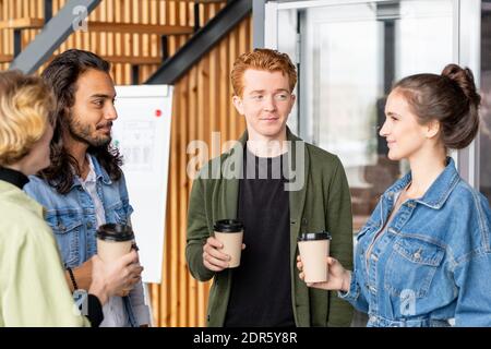 Gruppo di giovani direttori d'ufficio interculturali o creativi in casualwear che beve e discute di nuove idee alla pausa caffè Foto Stock