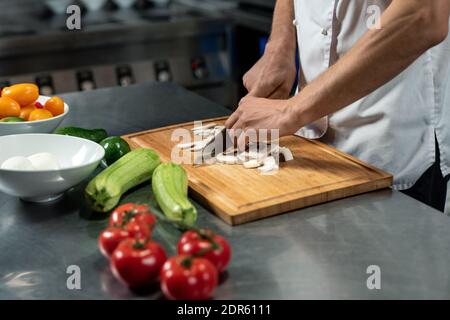 Mani di giovane chef maschile del ristorante moderno in bianco uniforme in piedi da grande tavolo in cucina e taglio funghi su tavola di legno Foto Stock