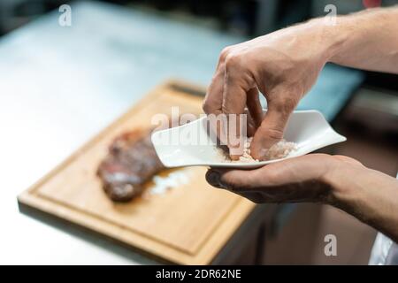 Mani di chef che prendono il sale marino per aggiungerlo a. bistecca di manzo arrosto su tavola di legno mentre si sta in piedi al tavolo e preparare il pasto per il cliente Foto Stock