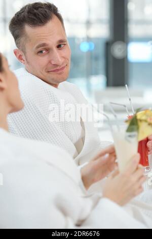 Felice giovane uomo riposante in accappatoio bianco guardando il suo moglie durante la conversazione mentre entrambi si siedono su sdraio e avere cocktail Foto Stock