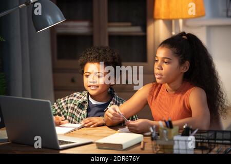 Due simpatici fratelli di etnia africana seduti al tavolo davanti al laptop e partecipando a lezioni online mentre ascoltare la spiegazione dell'insegnante Foto Stock